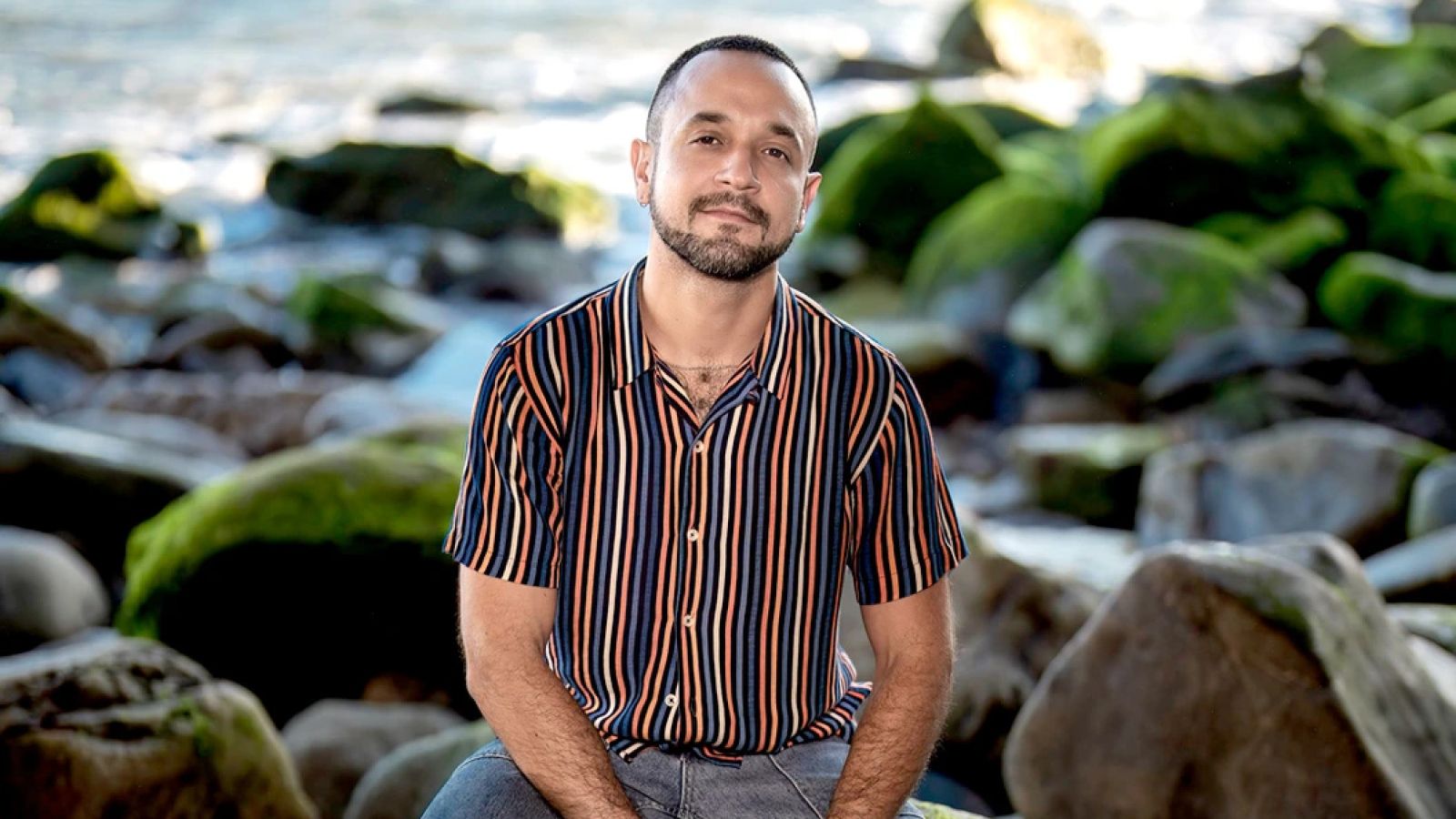 Fernando Morillo smiling while sitting on rocks by the beach with the ocean behind him.