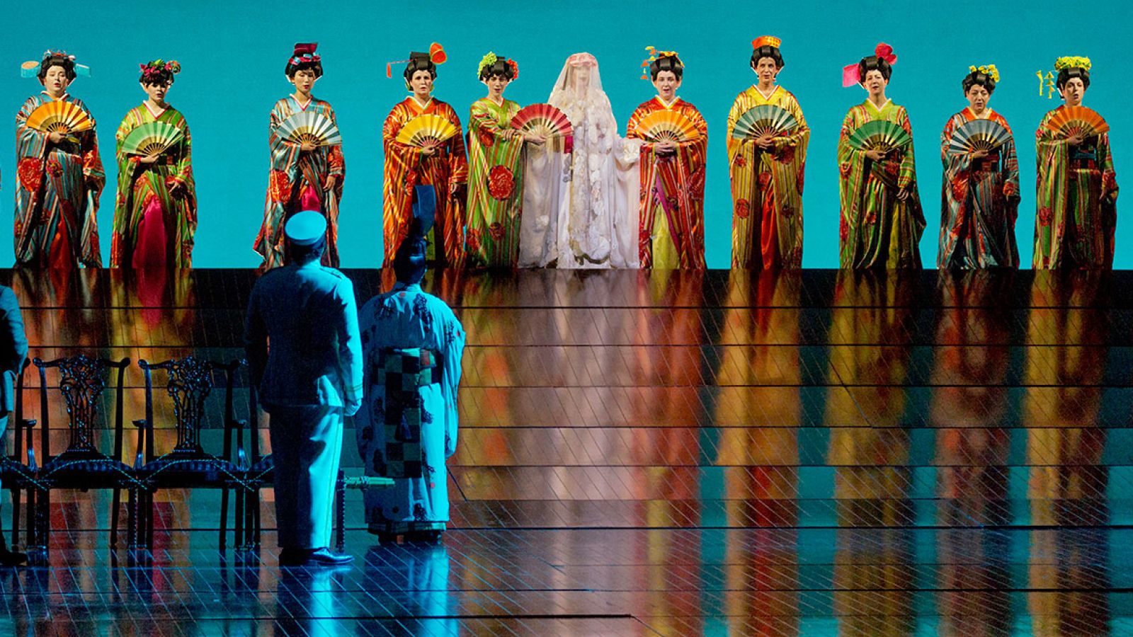 A bride stands in the center of a line of women. Two men stand facing the women with their back to the stage.