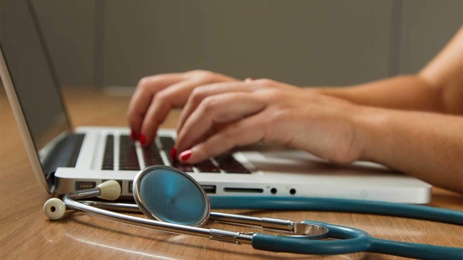 Photo of person using laptop with a stethoscope nearby.