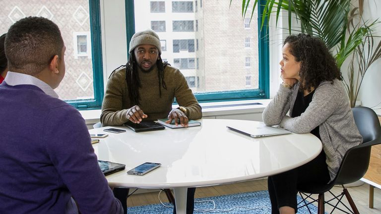 Photo - Three people sitting at an office table listen to a young man.