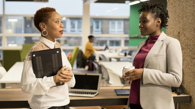 Two Black women are talking in an office with a laptop and tablet behind them.