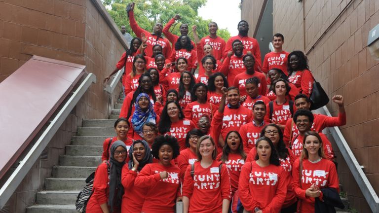 A group of students smile and wave while wearing matching red CUNY Cultural Corps shirts.