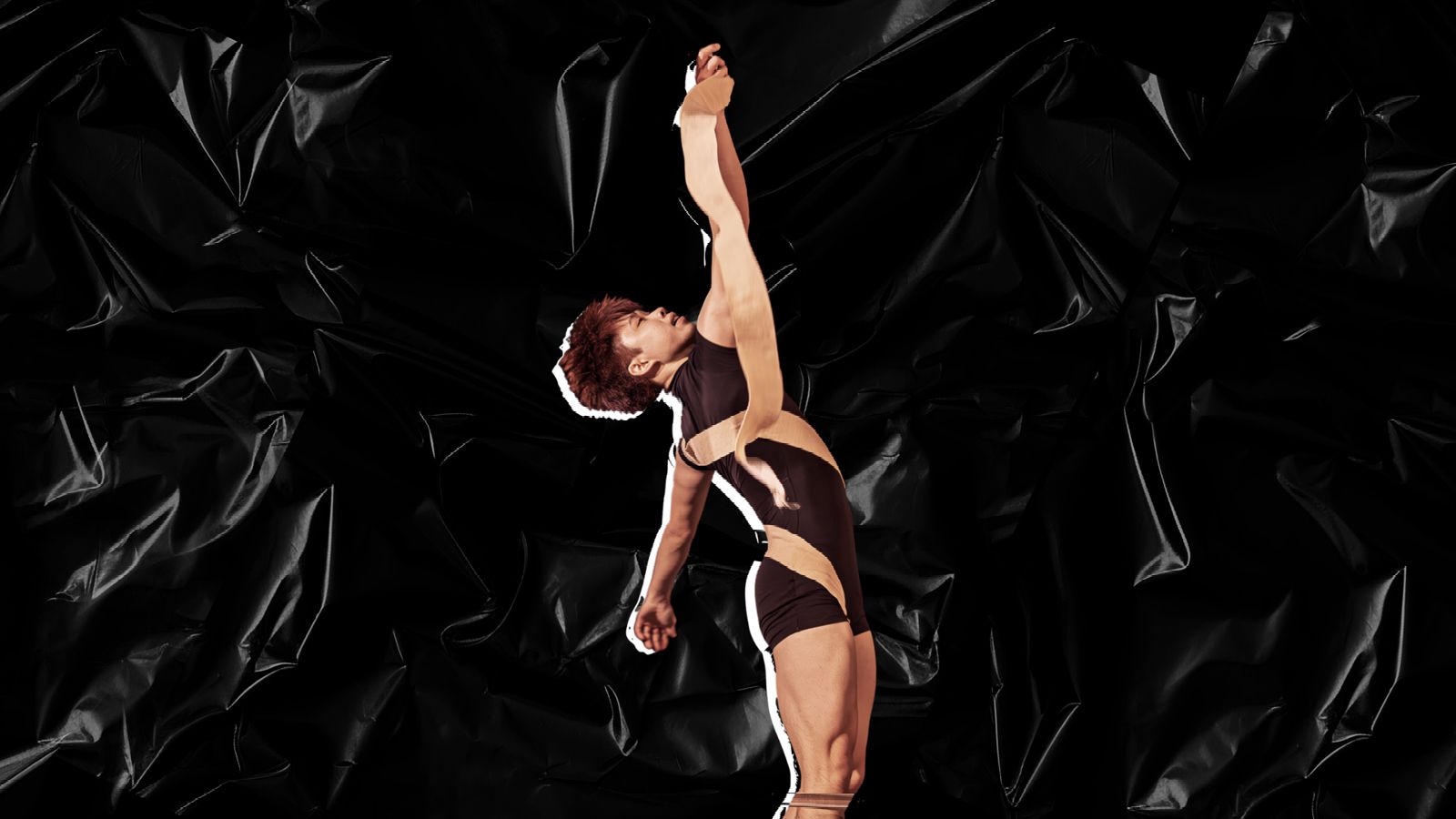 Dancer with her arms in the air holding a tan ribbon in front of a black background.