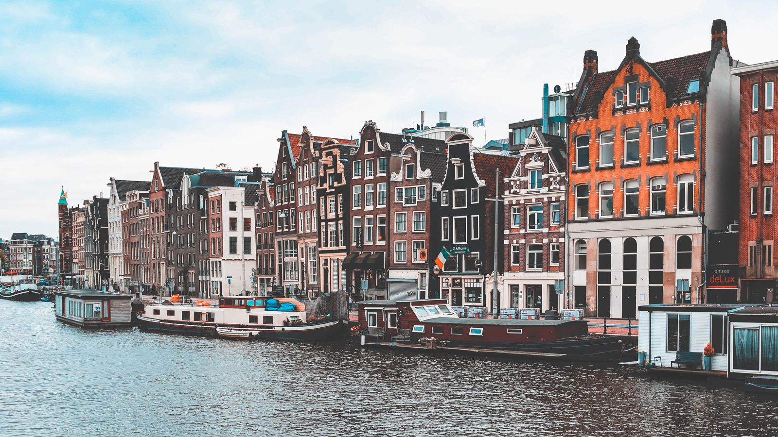 Photo: Boats and narrow buildings line a canal under a bright sky in Amsterdam, Netherlands.