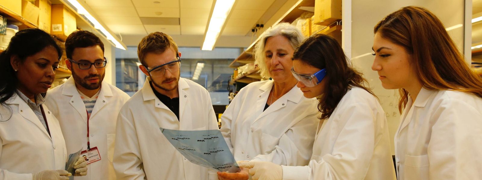 Students and Faculty wearing lab gowns reading results of their experiment.