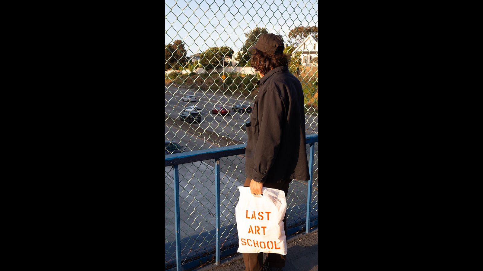 Person standing on overpass carrying a tote that says "Last Art School"