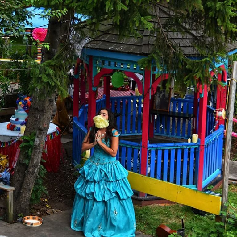 Actress is bright blue dress smells a bouquet in front of a brightly colored Gazebo.