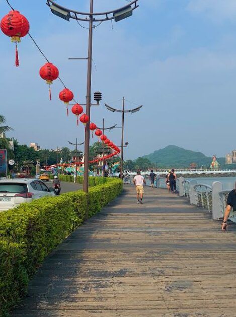 a person walking away from the viewer down a boardwalk near a river, with a row of red lanterns