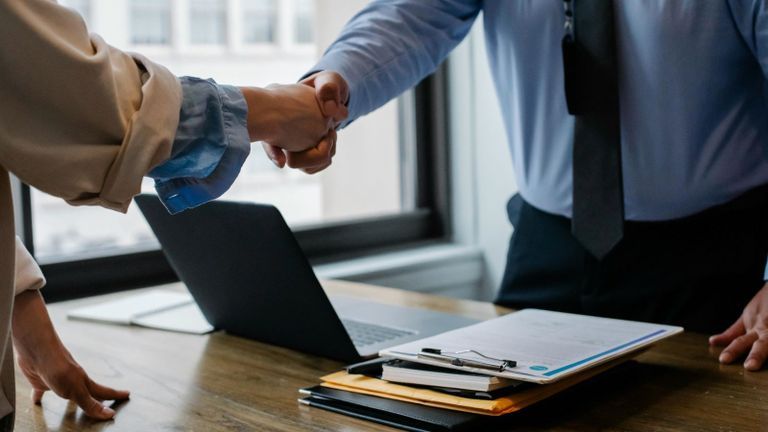 Two people shaking hands over a desk with a laptop and documents in an office setting.