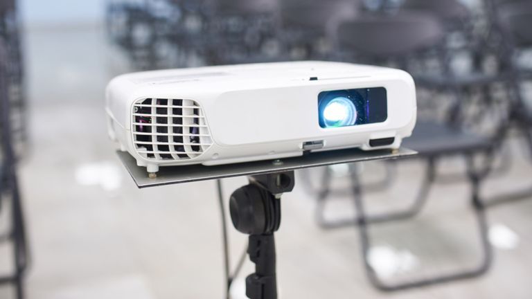 A white projector on top of a black stand in the middle of a classroom, with empty chairs in the background.