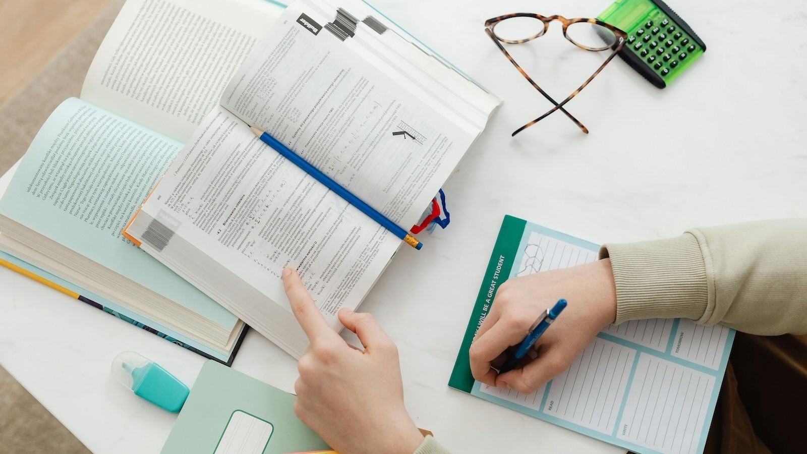 Overhead perspective of a student’s hands taking notes in a book at a desk, with study materials scattered around.
