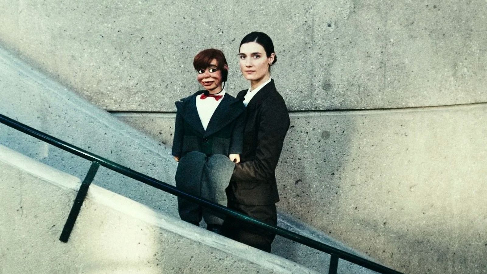 Sophie Becker standing on a staircase holding a ventriloquist dummy, dressed in matching suits.