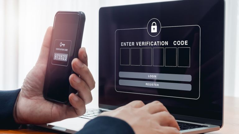 Person at laptop and holding phone, entering verification code for MFA