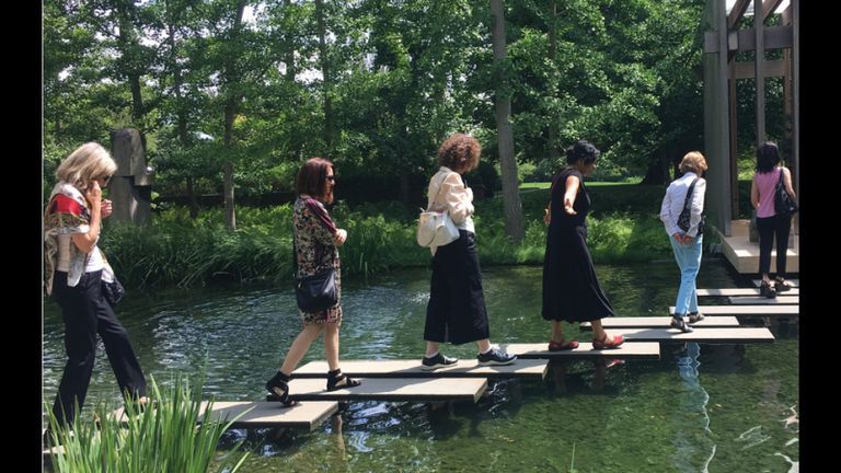Women walk across square rocks in a pond.
