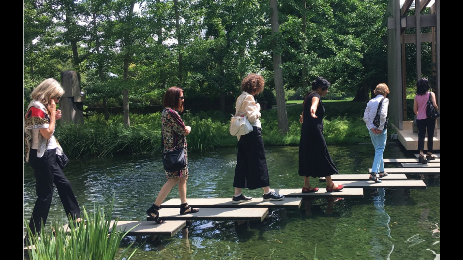 Women walk across square rocks in a pond.
