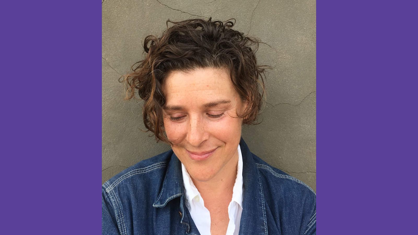 Photo of Zoe Leonard wearing a white dress shirt and jean jacket smiling and looking down.